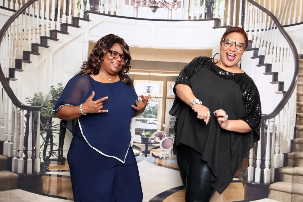 Southern Gals hosts Carmen and Felicia posing together in front of a grand spiral staircase